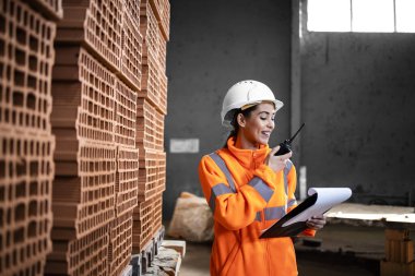 Kadın fabrika işçisi inşaat endüstrisi için kil tuğla yığınının yanında duruyor ve radyo envanterini kontrol ediyor..