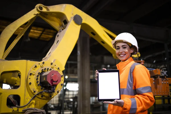 Female factory engineer holding tablet computer and standing by the industrial robotic manufacturing machine.
