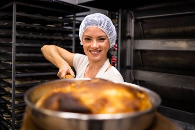 Çalışkan kadın fırıncının portresi fırının yanında duruyor ve tepsiyi yemek üretim fırınında taze pişmiş turtayla tutuyor..