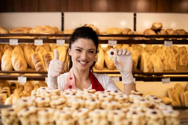 Portrait of confectioner person holding freshly baked cookies and thumbs up in bakery shop.