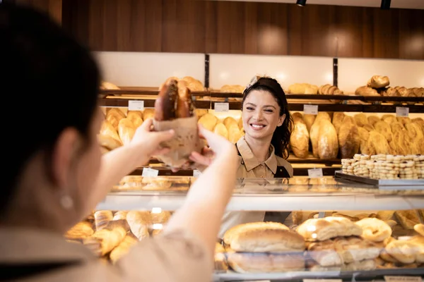 Beautiful smiling bakery worker selling fresh pastry to the customer in bakery shop.