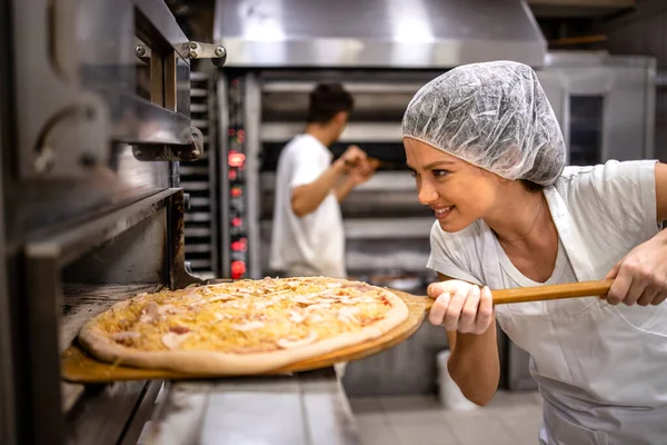 Working in pizza restaurant. Female chef in white uniform and hairnet putting pizza in the oven for baking process.
