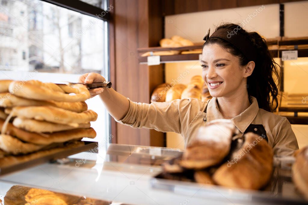 Beautiful smiling bakery worker selling fresh pastry to the customer in bakery shop.
