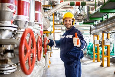 Isıtma fabrikası mühendisinin portresi doğalgaz boru hatlarının yanında duruyor ve başparmak kaldırıyor..