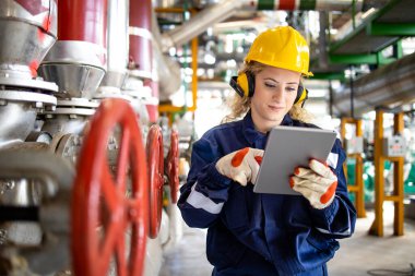 Rafineri işçisi, baret ve kulaklıklı. Petrol ve gaz üretimi için tablet bilgisayarında parametreleri kontrol ediyor..