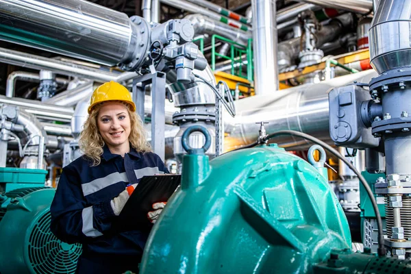 Portrait of female industrial or refinery worker standing by generators and gas pipes and holding notebook.