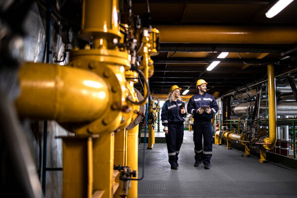 Industrial workers in protective work wear walking by gas pipeline inside refinery and checking oil production.