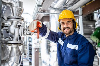 Portrait of smiling bearded refinery engineer or worker standing by pipes in oil production industry.