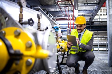 Production manager in elegant suit and reflective vest checking gas production in refinery or power plant.
