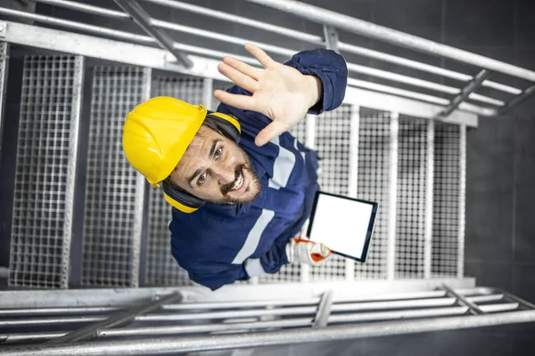 Top view of smiling caucasian worker wearing protective uniform and helmet waving to the camera in factory.