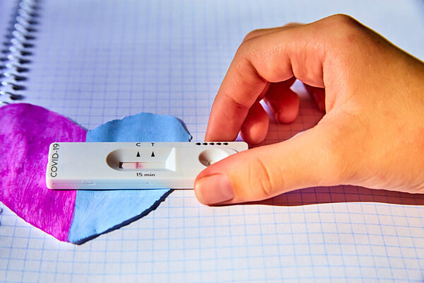 children's hand holds Fast and simple Self-Test COVID-19 Antigen with negative result. he provides results in 15 minutes. using nasal swab.on hand-carved violet-blue heart and checkered school leaflet