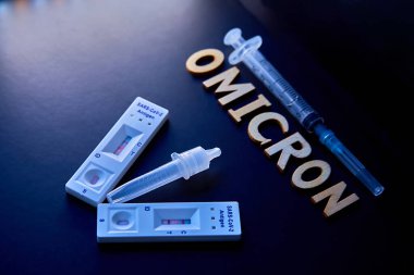 Omicron kelimesi ahşap harfler, şırınga, tıbbi enjeksiyon şişesi ve 2 covid-19 antijen testinden pozitif sonuç çıktı. Konsept-mutasyona uğramış koronavirüs SARS-CoV-2 grip salgını.