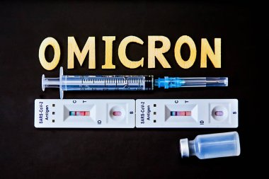 Omicron kelimesi ahşap harfler, şırınga, tıbbi enjeksiyon şişesi ve 2 covid-19 antijen testinden pozitif sonuç çıktı. Konsept-mutasyona uğramış koronavirüs SARS-CoV-2 grip salgını.