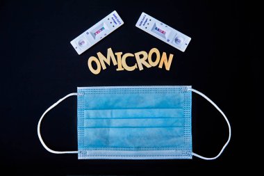 Omicron kelimesi ahşap harflerden, mavi tıbbi maskeden ve 2 covid-19 antijen testinden yapılmış. Siyah arka plan pozitif. Tıbbi konsept - Mutasyona uğramış koronavirüs SARS-CoV-2 grip salgını