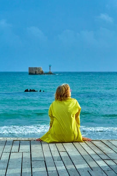 Woman Bright Yellow Shirt Sits Her Back Seashore Looks Distance — Stock Photo, Image