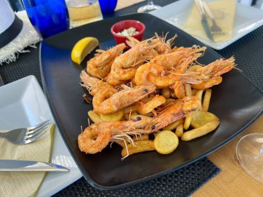 Izgara büyük kral karidesleri siyah bir tabakta patates kızartmasının üzerinde bir dilim limon ve sos ile uzanır.