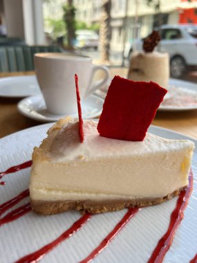 Ahududu ya da çilek reçeli serpilmiş beyaz bir tabakta beyaz bir fincan kahvenin yanında cheesecake.