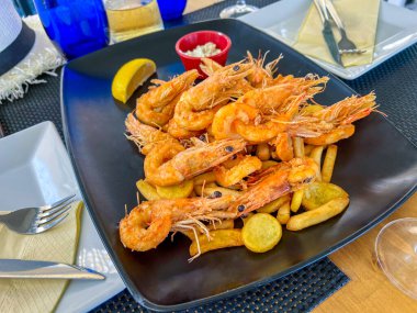 Izgara büyük kral karidesleri siyah bir tabakta patates kızartmasının üzerinde bir dilim limon ve sos ile uzanır.