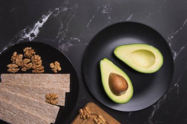 Siyah tabakta iki avokado parçası, görünür tohumla ikiye bölünmüş ve koyu arkaplanda İngiliz cevizleri var.