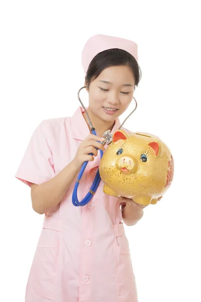 Beautiful Asian nurse pretending to listen to the heart beat of a piggy bank isolated on a white background.  Illustrating the high cost of medical care — Stok fotoğraf