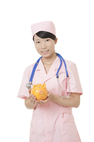 Linda enfermeira asiática segurando um banco porquinho isolado em um fundo branco. Ilustrando o alto custo da assistência médica — Fotografia de Stock
