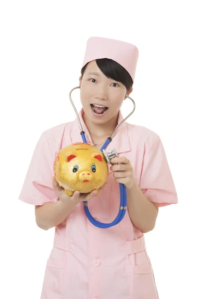 Beautiful Asian nurse pretending to listen to the heart beat of a piggy bank isolated on a white background.  Illustrating the high cost of medical care — Φωτογραφία Αρχείου