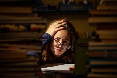 Gözlüklü stresli bir kadın final sınavları için kütüphanede ders çalışıyor..