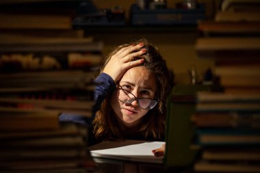 A stressed woman with glasses studying in a library for her final exams. clipart
