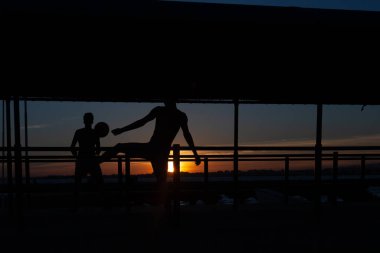 Gün batımında Portekiz 'de bir sahil yolunda futbol oynayan iki gencin silüeti.