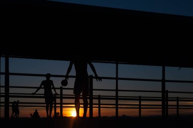 Gün batımında Portekiz 'de bir sahil yolunda futbol oynayan iki gencin silüeti.