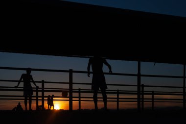 Gün batımında Portekiz 'de bir sahil yolunda futbol oynayan iki gencin silüeti.