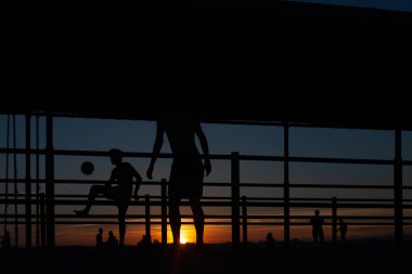 Gün batımında Portekiz 'de bir sahil yolunda futbol oynayan iki gencin silüeti.