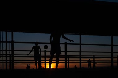 Gün batımında Portekiz 'de bir sahil yolunda futbol oynayan iki gencin silüeti.