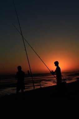 Gün batımında sahilde iki balıkçının silueti.