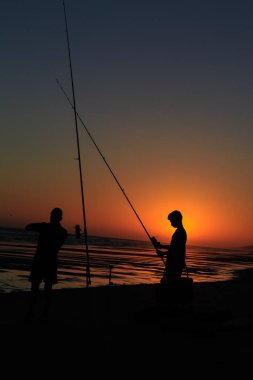 Gün batımında sahilde iki balıkçının silueti.