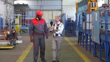 Kafkas Üretim Müdürü ve Koruyucu İş Giyimi 'nde Siyah İşçi Bir Fabrika Salonunda Konuşuyor ve Yürüyor. Denetmen dijital tablet kullanıyor ve bir işçiyle üretim sürecini tartışıyor.