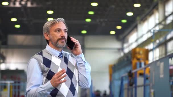 在工厂里讲智能手机的成熟经理 在工业室内环境中表现出自信 灰白头发的商人形象 在工厂里站着用智能手机的成熟男人 — 图库视频影像