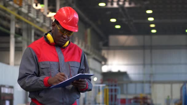 年轻的非洲裔美国工人在一家工厂的剪贴板上书写个人防护设备 穿红色镜片 黄色护目镜和工作服的黑人产业工人的蜡像 — 图库视频影像