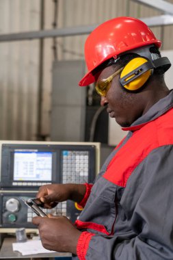 Afrika kökenli Amerikan CNC Makine Operatörü Kişisel Koruyucu Ekipman Ölçülerini Dijital Vernier Caliper ile kontrol ediyor. Kırmızı Miğferli Siyah Endüstriyel İşçi Portresi, Sarı Güvenlik Gözlüğü, İşitme Koruyucuları ve Fabrikada İş Üniforması.
