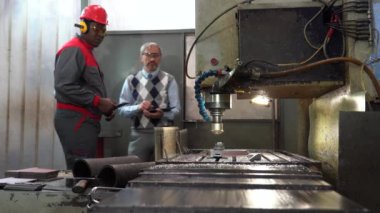 Afro-Amerikan CNC makine operatörü ve Kafkas kalite kontrol müfettişi bir değirmen makinesinin işini kontrol ediyor. CNC Değirmen Makinesi Kesme Çeliği. Çok Irklı İş Arkadaşları Konuşuyor.