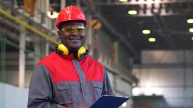 Koruyucu İş Giyimi 'nde gülümseyen Afrikalı genç bir işçi pano tutuyor ve başparmağını kaldırıyor. Kırmızı Miğferli, Güvenlik Gözlüklü ve İş Üniformalı Siyah Endüstriyel İşçi Portresi.