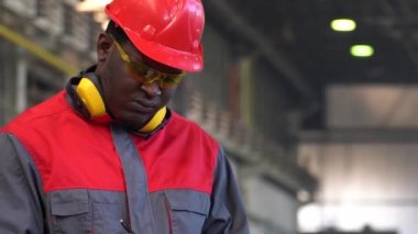 Afrikalı Amerikalı Koruyucu İş Giyimi İşçisi bir fabrikadaki panoya yazı yazıyor. Kırmızı Miğferli, Sarı Güvenlik Gözlüklü ve İş Üniformalı Siyah Endüstriyel İşçi Portresi.