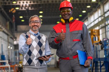 Smiling Black Worker in Protective Workwear And Caucasian Production Manager Giving Thumbs Up. Multiracial Industrial Co-Workers Looking At Camera And Showing Thumbs Up In A Factory.