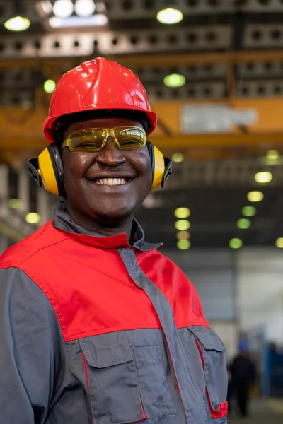 Smiling Black Worker In Personal Protective Equipment Looking At Camera. Portrait Of Industrial Worker In Red Helmet, Yellow Safety Goggles, Noise Reduction Earmuffs And Work Uniform In A Factory.