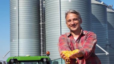 Happy Farmer, tahıl ambarına karşı yeni hasat edilmiş tahılları gösteriyor. Çiftçi, mısır tohumlarıyla dolu traktör römorkunda Tahıl ambarının önünde oturuyor. Etanol Biorefinery Siloları.