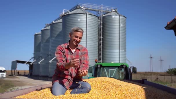 トラクタートレーラーに座っている幸せな農家穀物貯蔵ビンの前でトウモロコシの穀物の完全な 穀物サイロに対して収穫された穀物を示す笑顔の農家 収穫の時間だ とうもろこしの収量 — ストック動画
