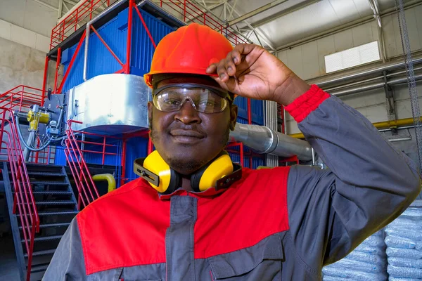 Portrait Of African American Worker In Protective Workwear In Central Boiler Plant. Black Worker In Red Helmet, Protective Eyewear, Hearing Protection Equipment And Work Uniform Looking At Camera.