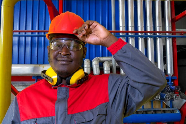 Portrait Of African American Worker In Protective Workwear In Industrial Interior. Blue-Collar Worker In Red Helmet, Protective Eyewear, Hearing Protection Equipment And Work Uniform Looking At Camera.