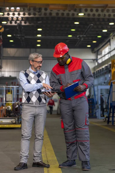 Production Manager With Digital Tablet Giving Instructions To Worker In Protective Workwear. Multiracial Industrial Co-Workers Standing In A Factory And Talking About Production Process. African American Worker Writing On Clipboard.