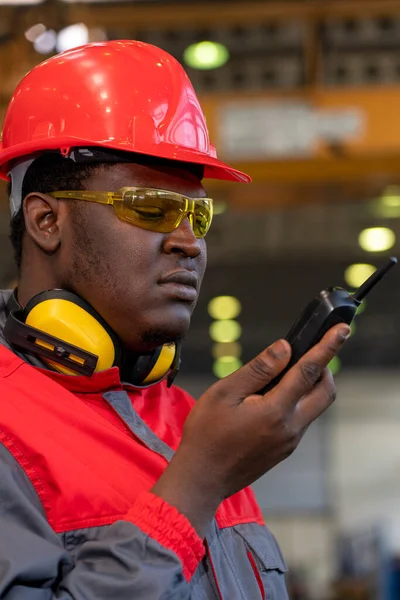 African American Worker In Protective Workwear Talking On Radio Communication Equipment In A Factory. Portrait Of Black Industrial Worker In Red Helmet, Yellow Safety Goggles, Hearing Protection Equipment And Work Uniform.